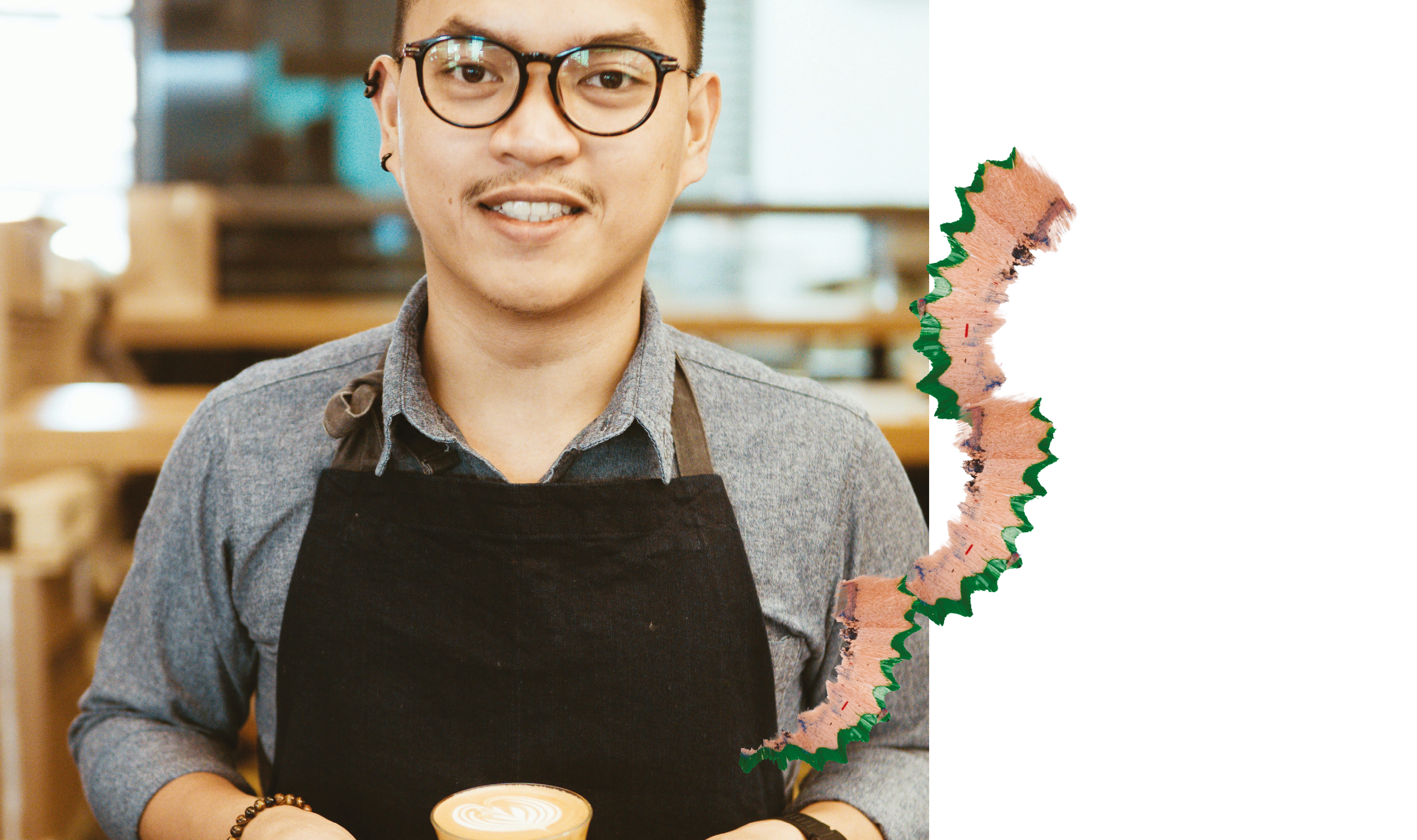 Ein Barista mit Brille und Schürze lächelt und hält eine Tasse Kaffee, daneben eine dekorative Bleistiftspäne.